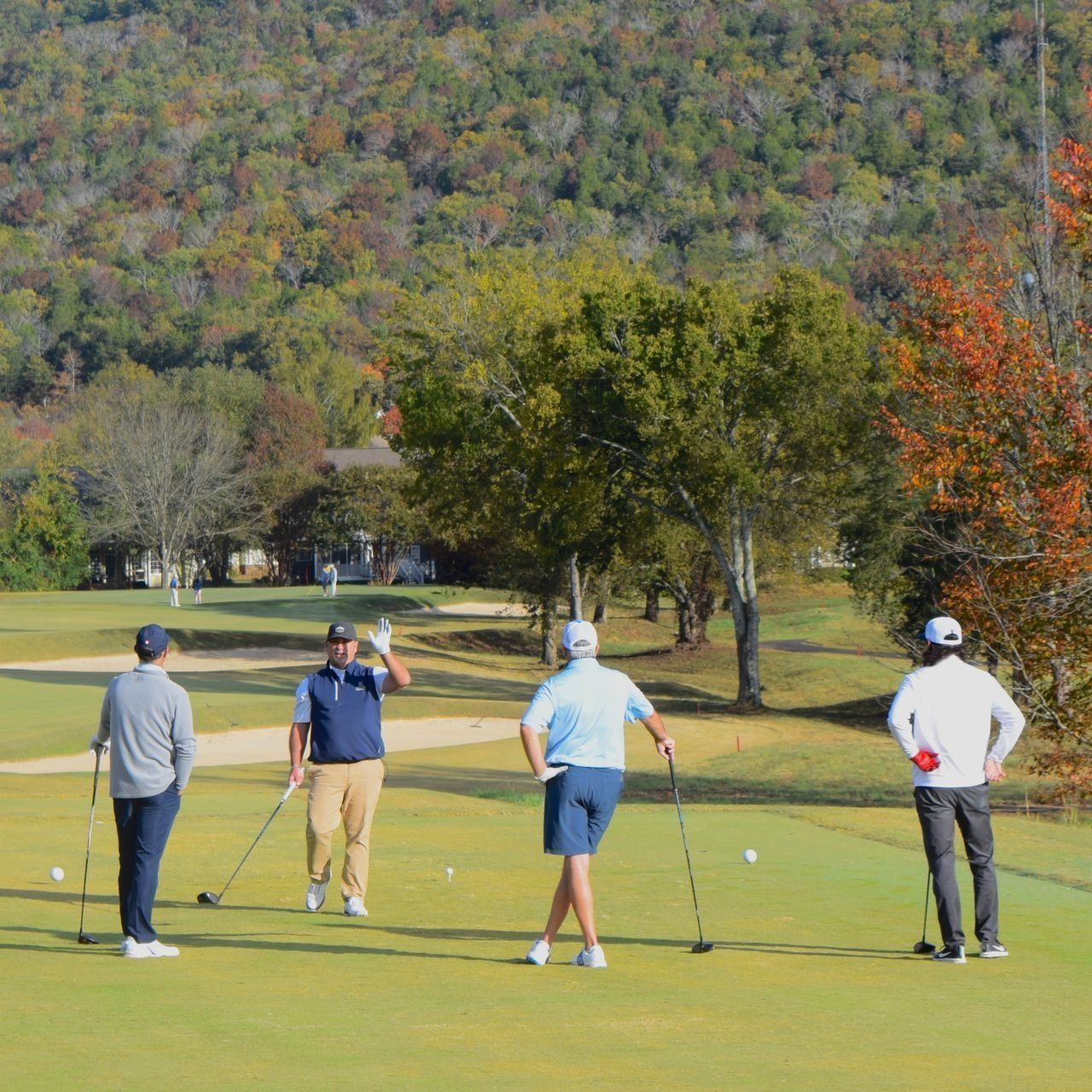 Four golfers standing on a green fairway, preparing to tee off with autumn trees and hills behind them