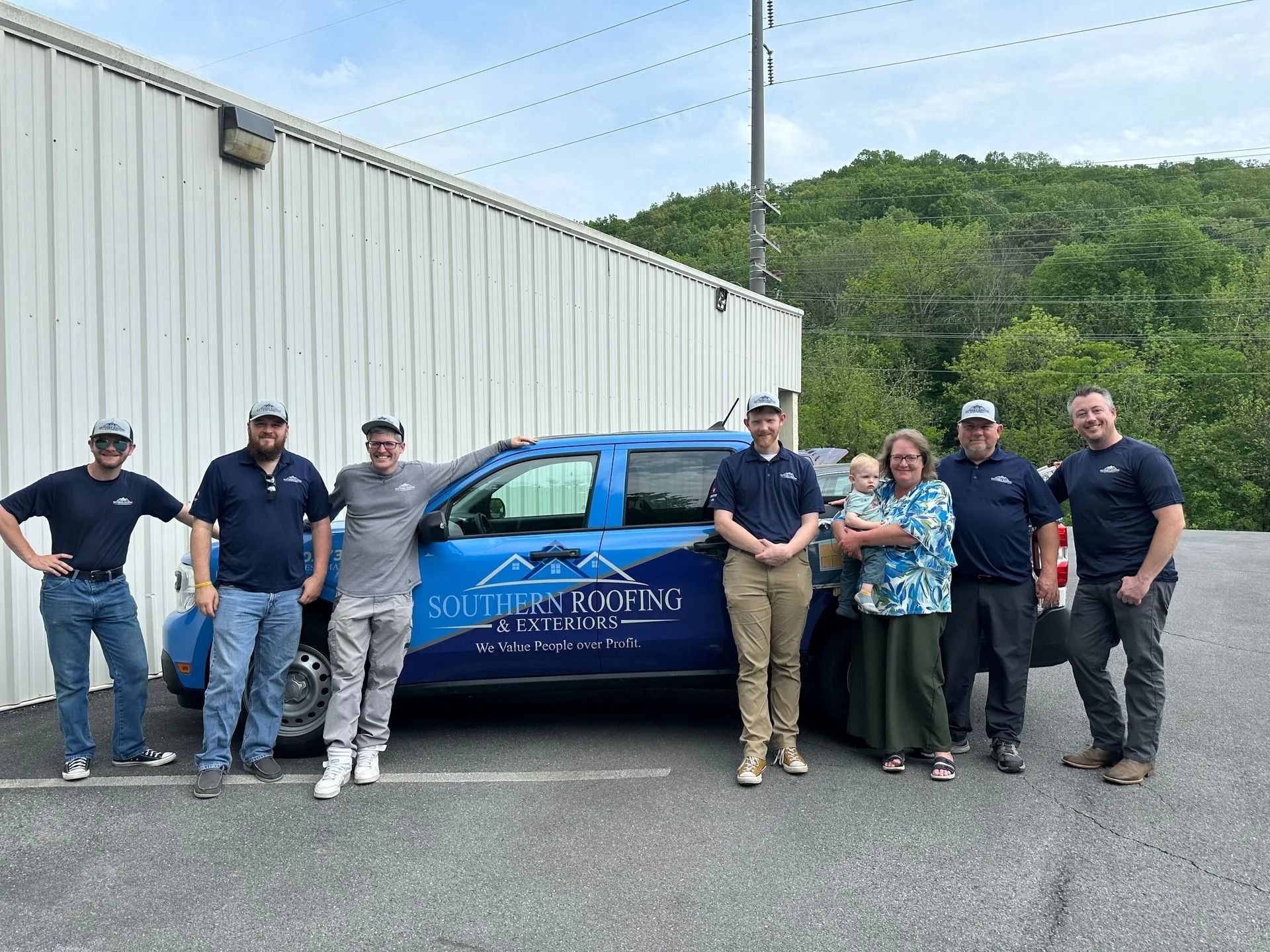 Six people posing beside a blue pickup truck in a parking lot near a metal building.