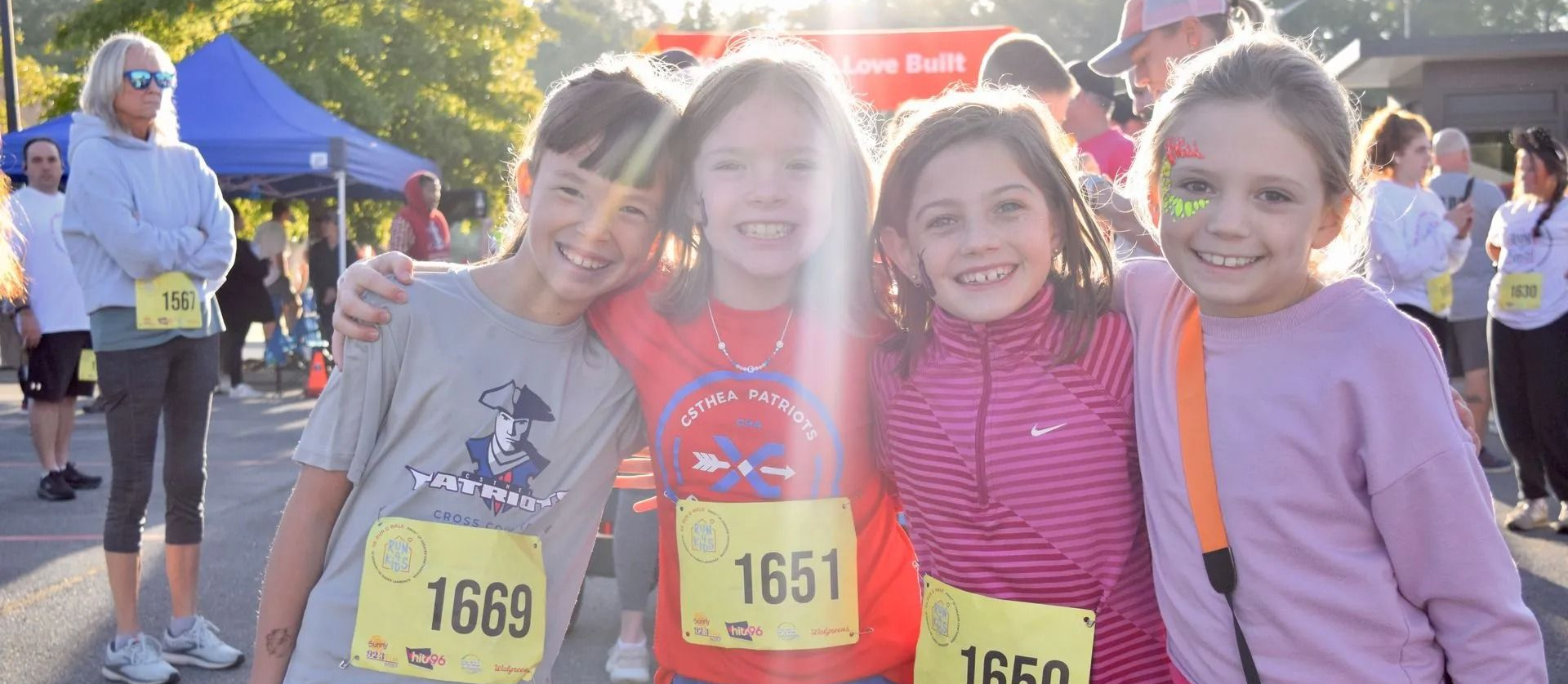Four smiling runners wearing bibs stand together at a race start, one in a pink striped shirt and orange suspenders.