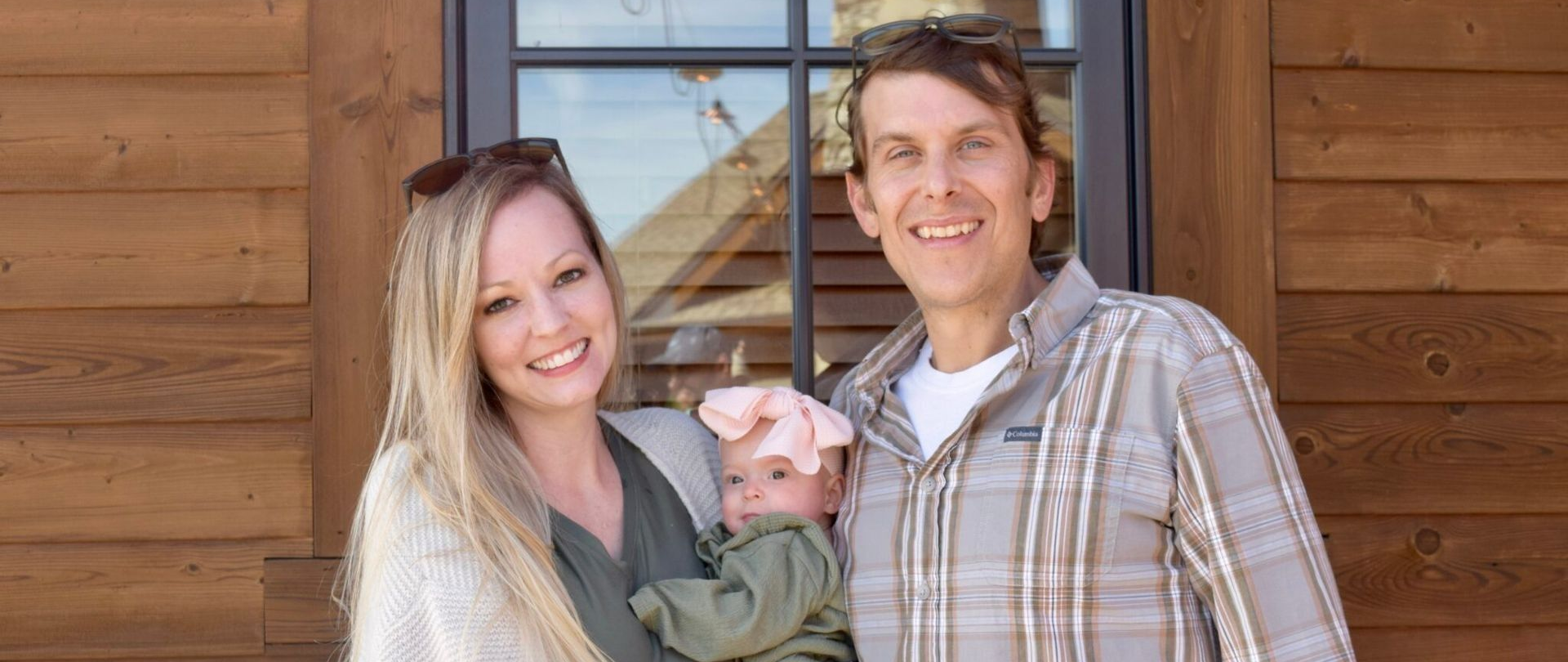 Smiling couple holding a baby in front of a wooden cabin wall