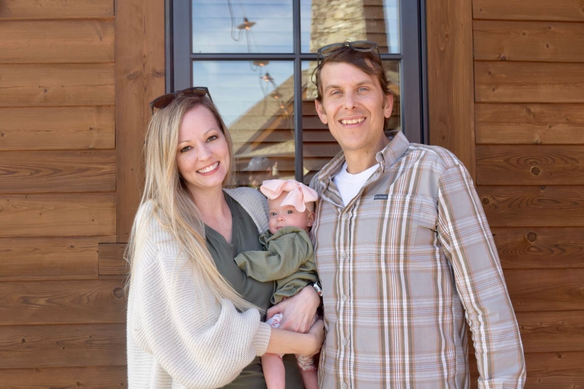Smiling couple holding a baby in front of a wooden building and window