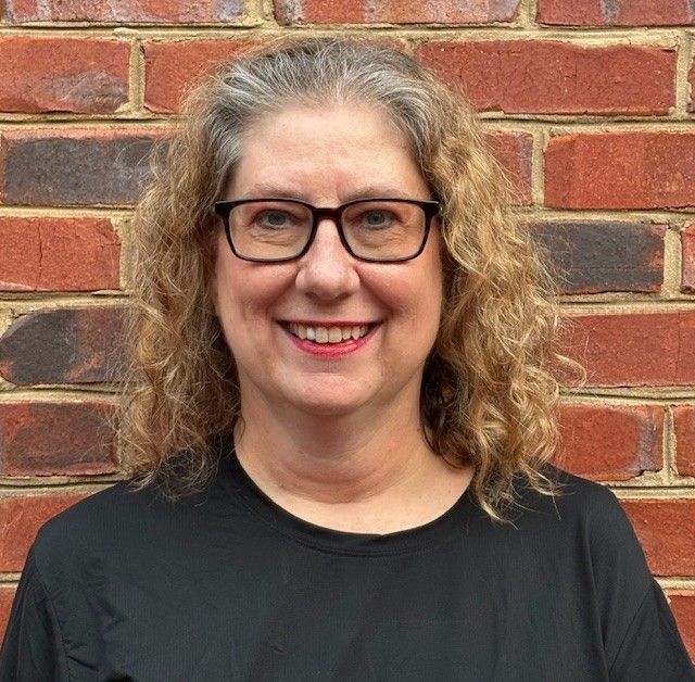 Smiling person with glasses in a black shirt standing against a red brick wall