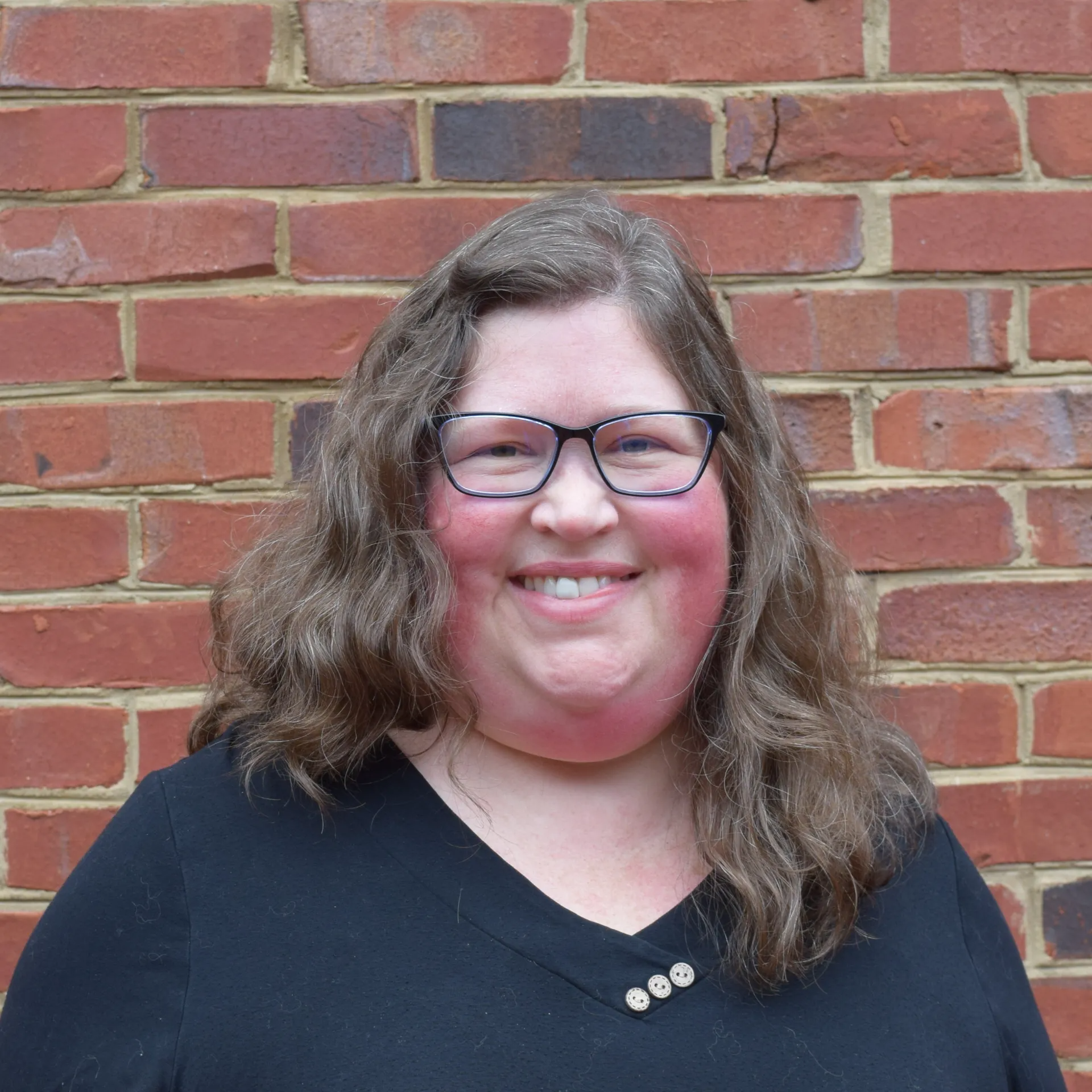 Smiling person with glasses and long hair standing against a red brick wall