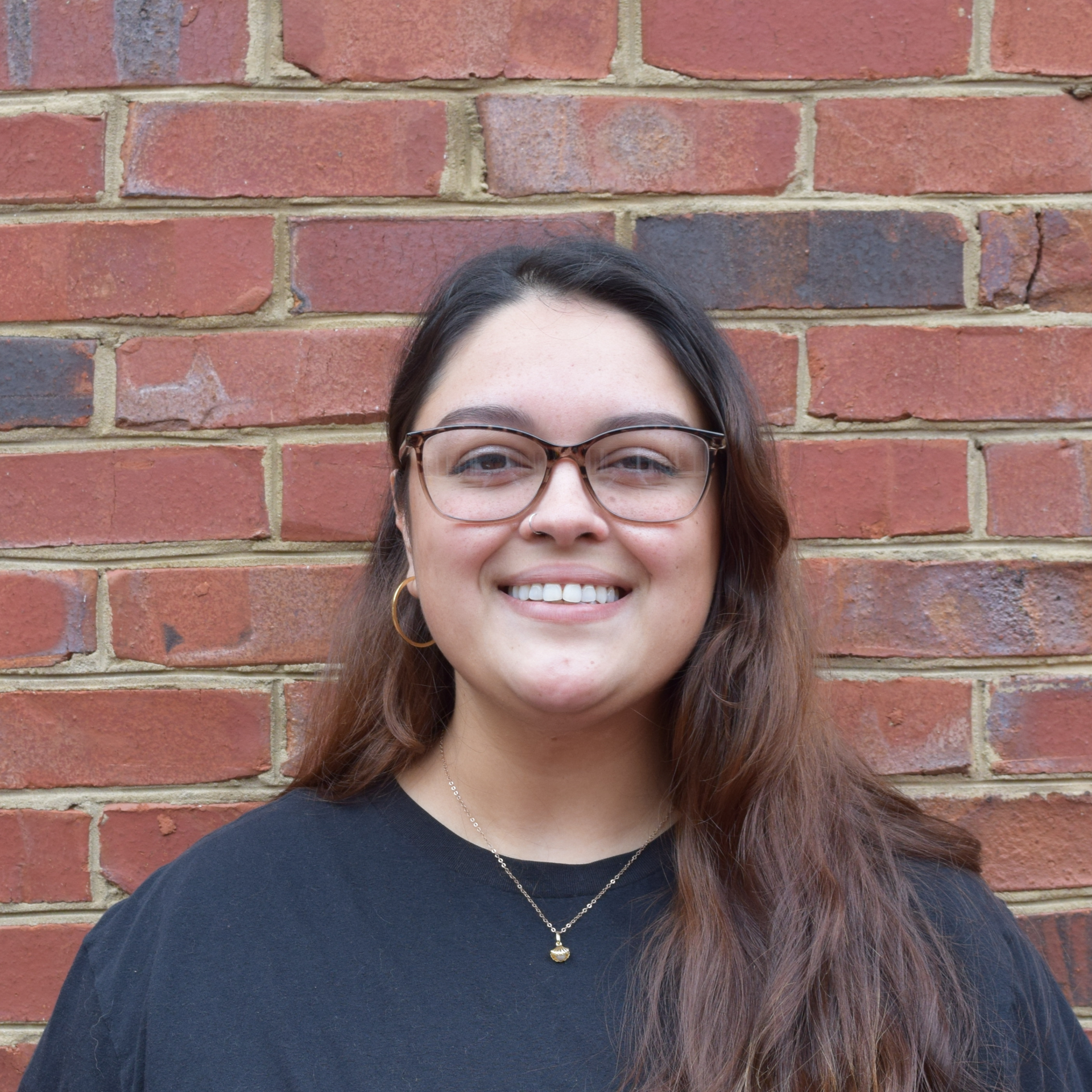 Smiling person with glasses in a black shirt standing against a red brick wall