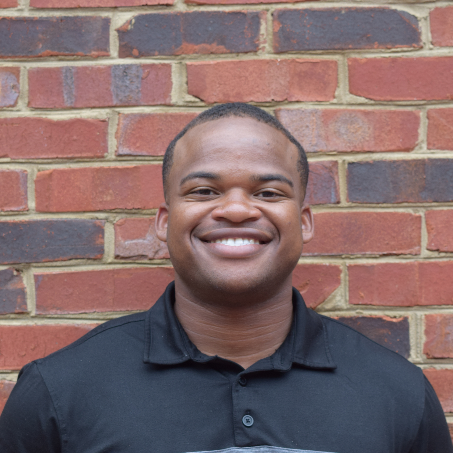 Smiling person in a black polo shirt standing against a red brick wall.