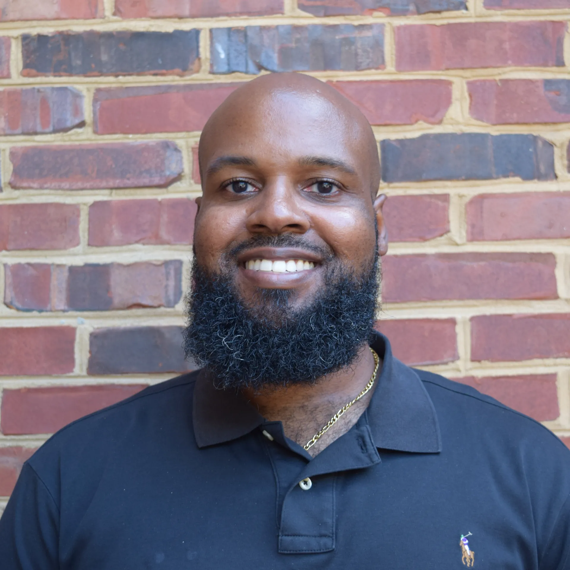 Smiling man with a beard wearing a navy polo shirt, standing in front of a brick wall