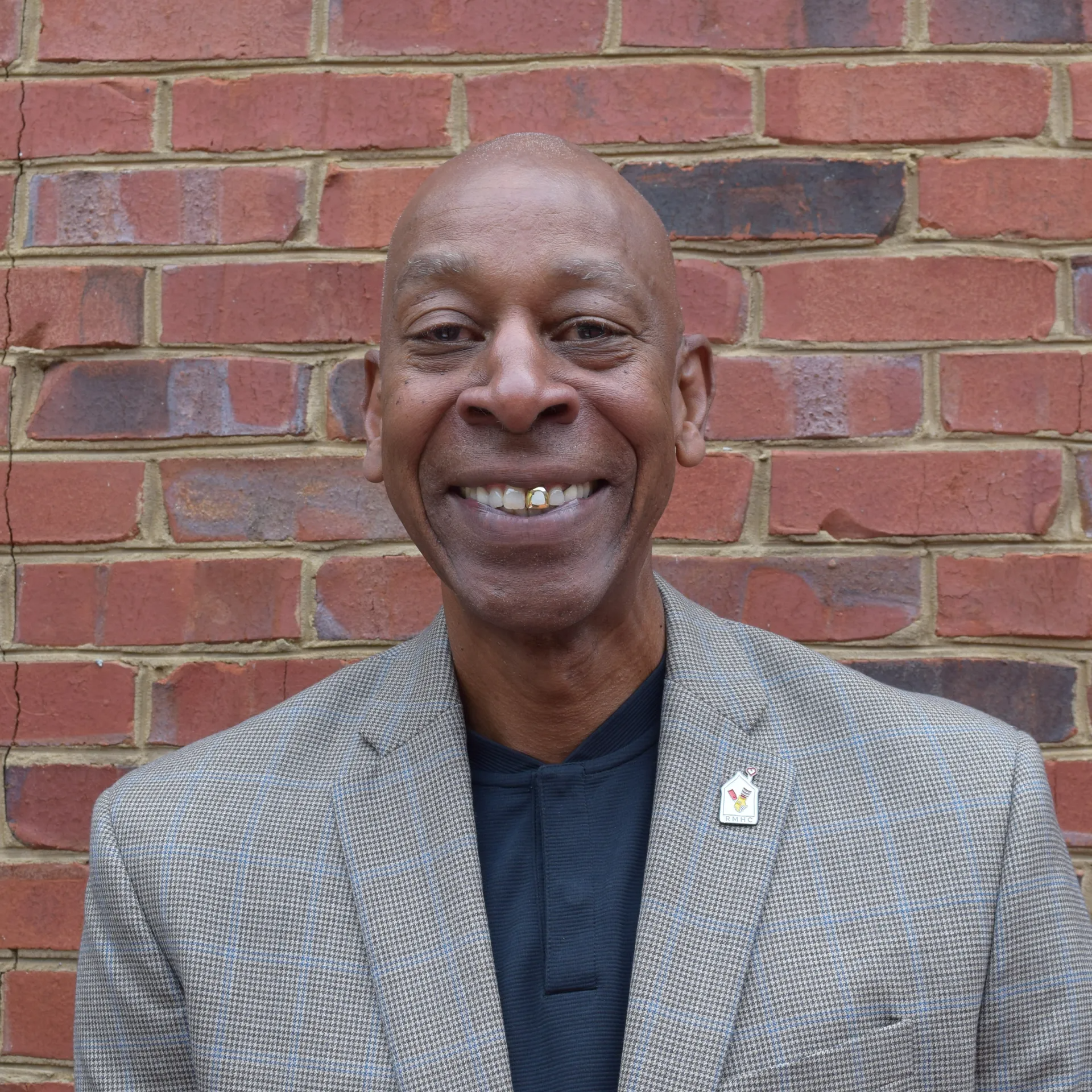 Smiling man in a gray blazer standing before a red brick wall