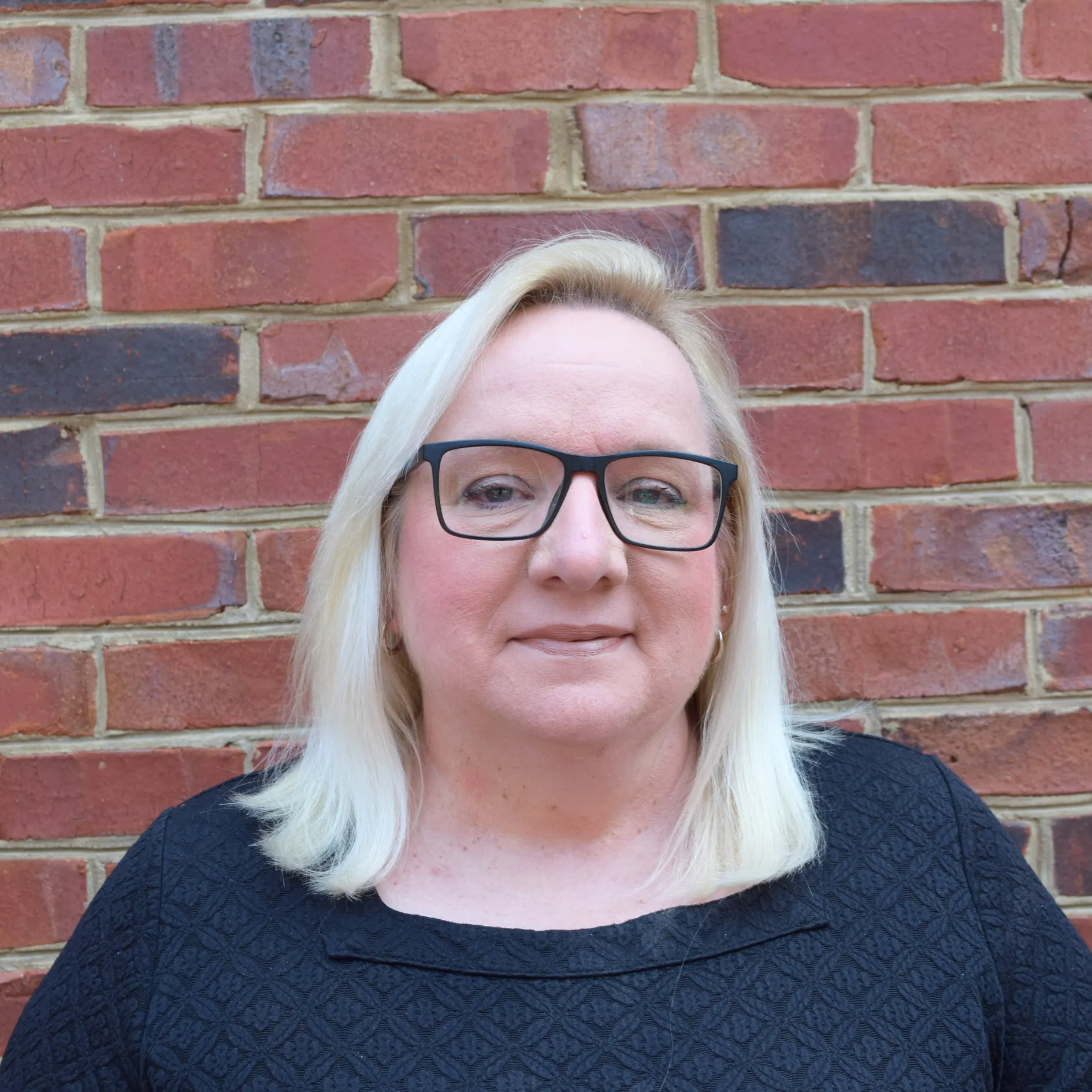 Portrait of a blonde person wearing glasses and a black top, standing against a red brick wall