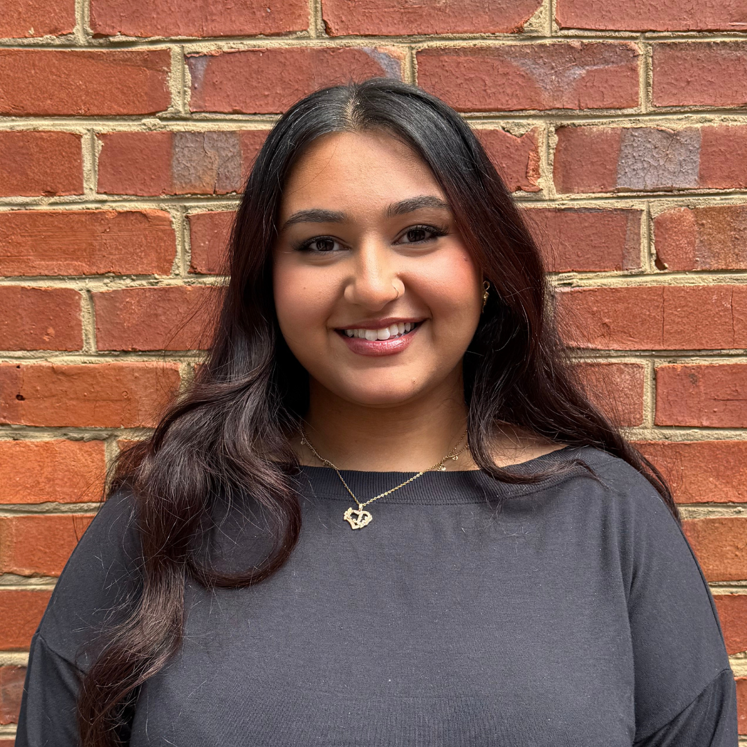Smiling person with long dark hair in a gray top, standing against a brick wall.