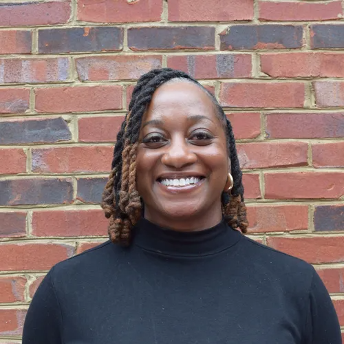 Smiling person in a black turtleneck standing against a brick wall.