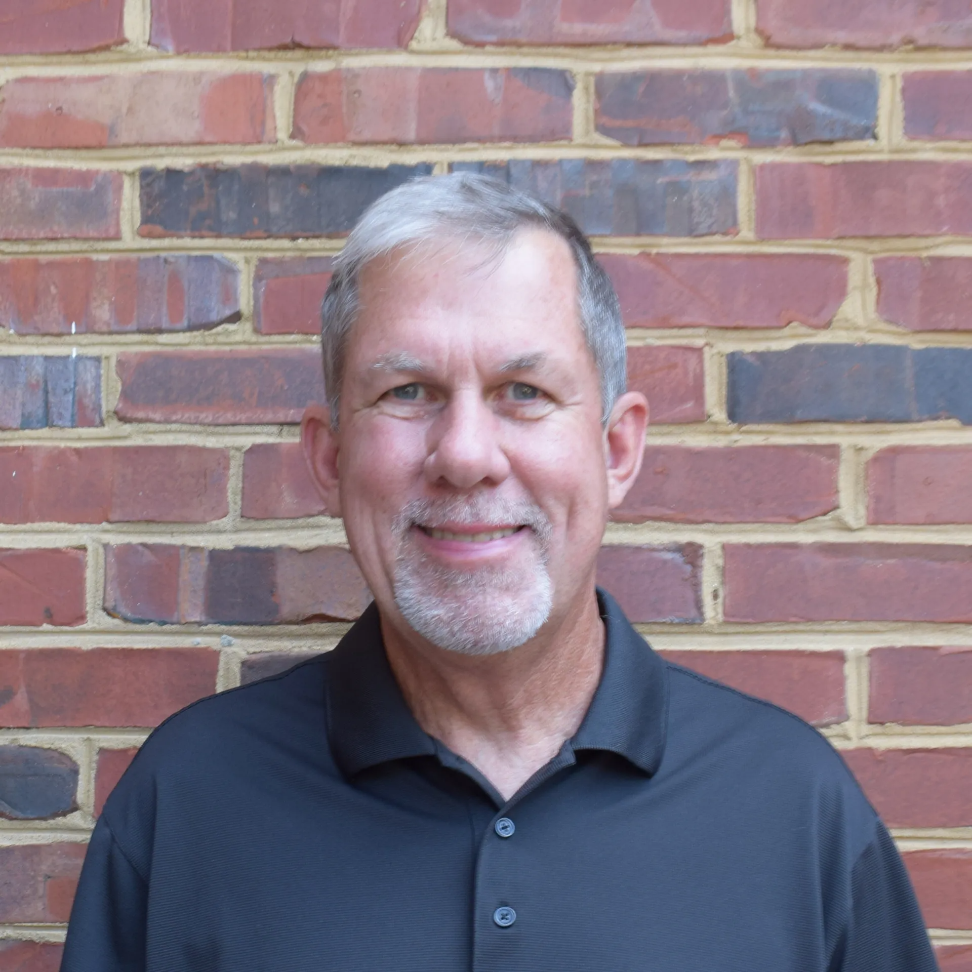 Smiling man in a dark polo shirt standing in front of a brick wall.