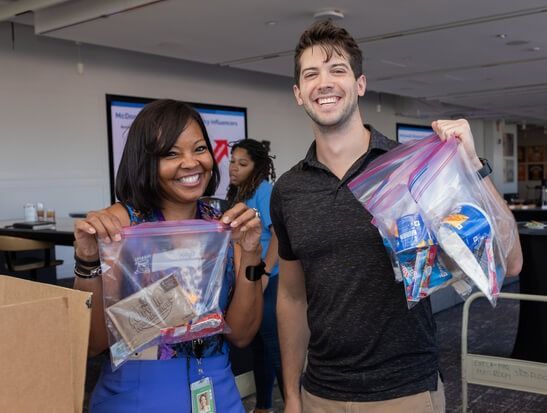 Two volunteers smiling and holding clear bags of donated items indoors.