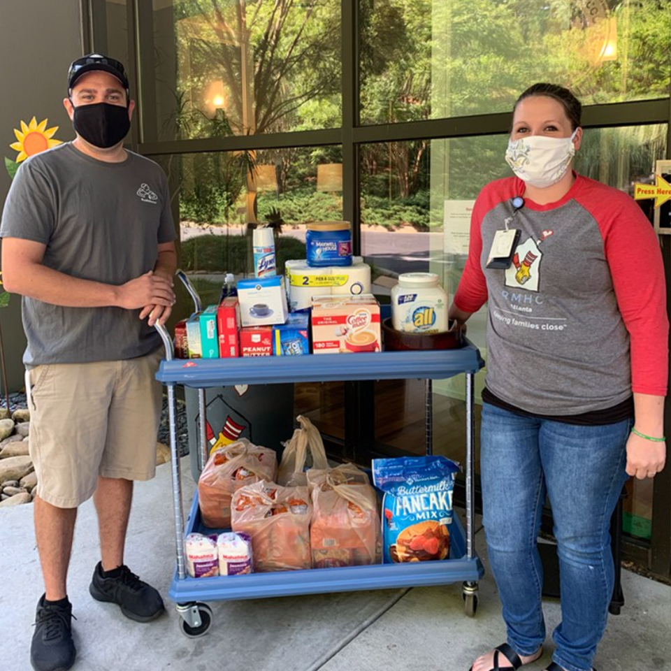 Two masked people stand beside a cart of donated groceries in a covered outdoor area.