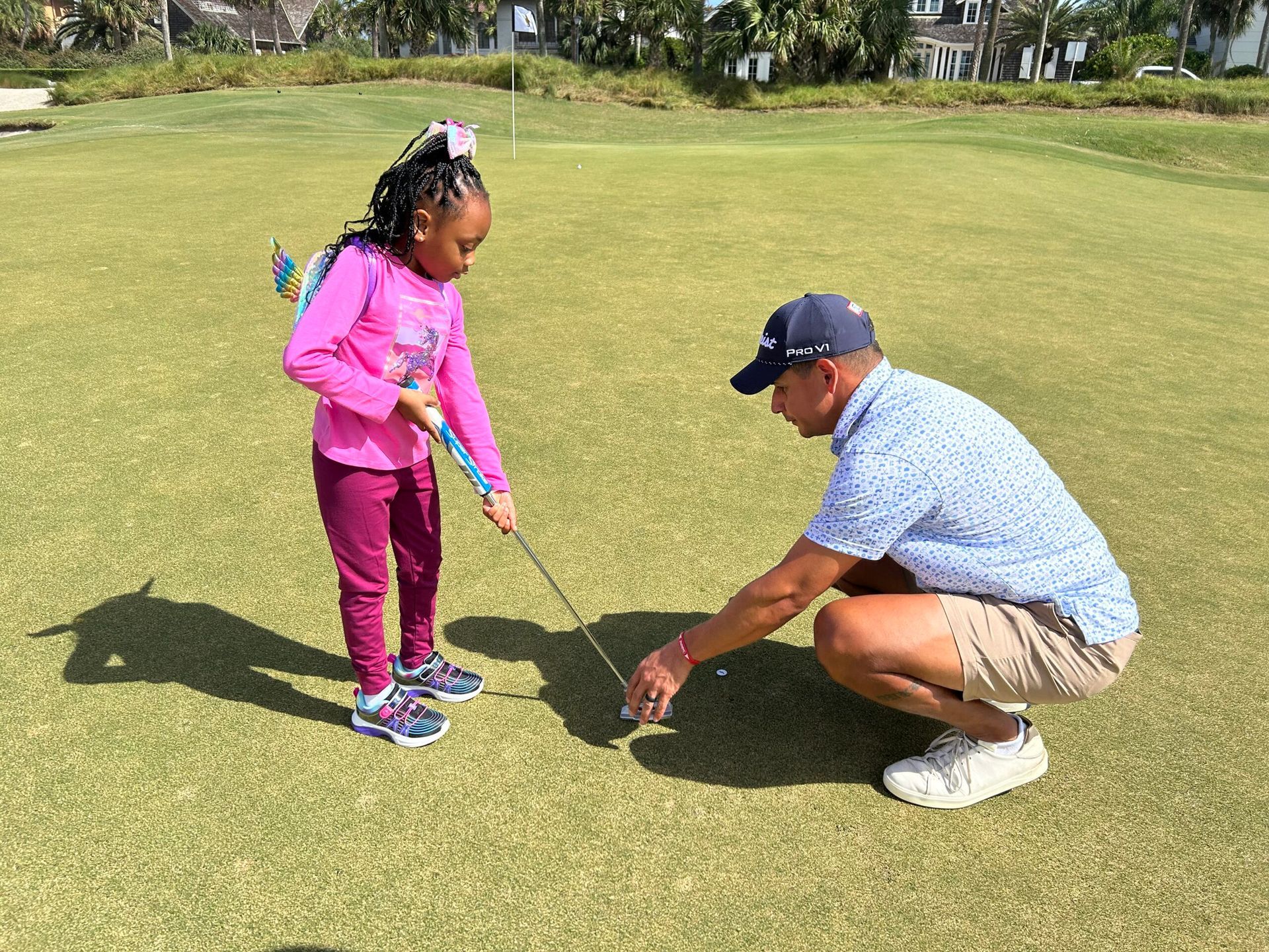 Adult kneels on a golf course, helping a child line up a putt with a club