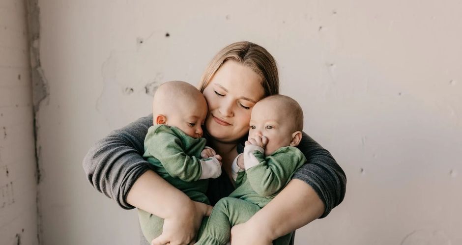 Person holding two babies in green outfits, smiling with eyes closed against a plain wall
