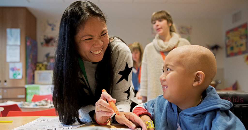 Teacher smiling at a child in a classroom, leaning over a table with school supplies.