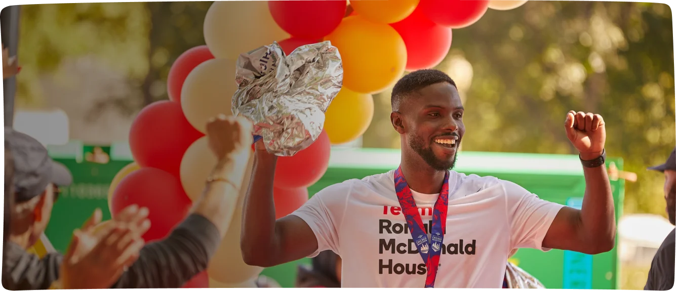 Man celebrating at a charity event with balloons and a gold medal, raising both hands and smiling