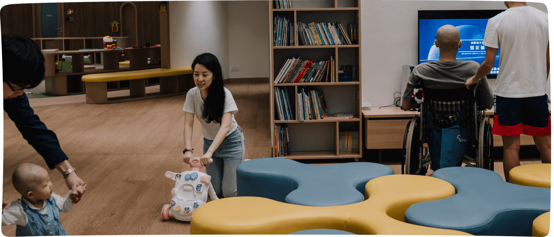 Children play in a library area with bookshelves, colorful seats, and adults nearby.