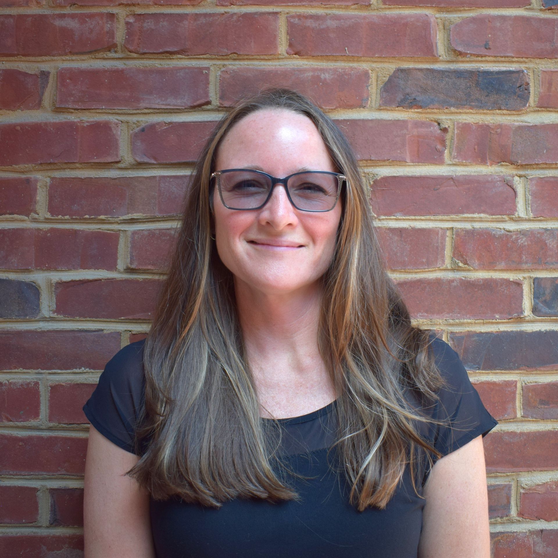 Person with glasses and long hair smiling against a red brick wall.