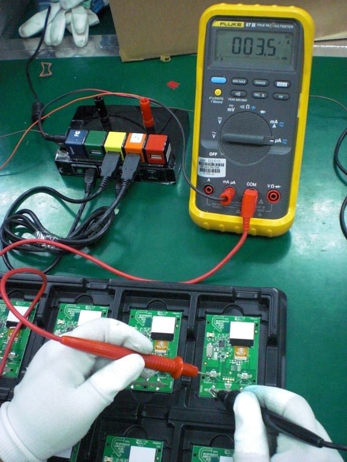A person wearing gloves tests a circuit board with a multimeter in a factory setting.