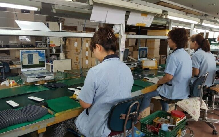 Factory workers assembling electronics at a workstation, with laptops and components in an industrial setting.