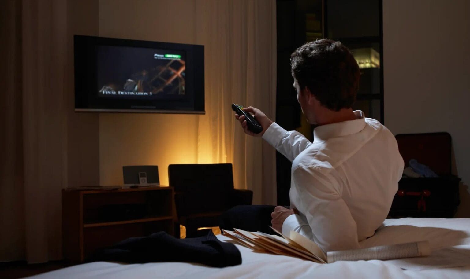 Man in white shirt on a bed, watching TV in a dimly lit hotel room, holding a remote.