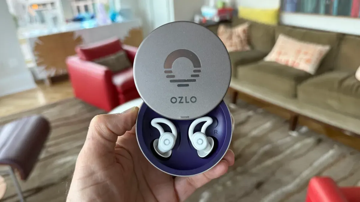 Person holding open charging case with two white sleep earbuds. The case is gray with purple interior.