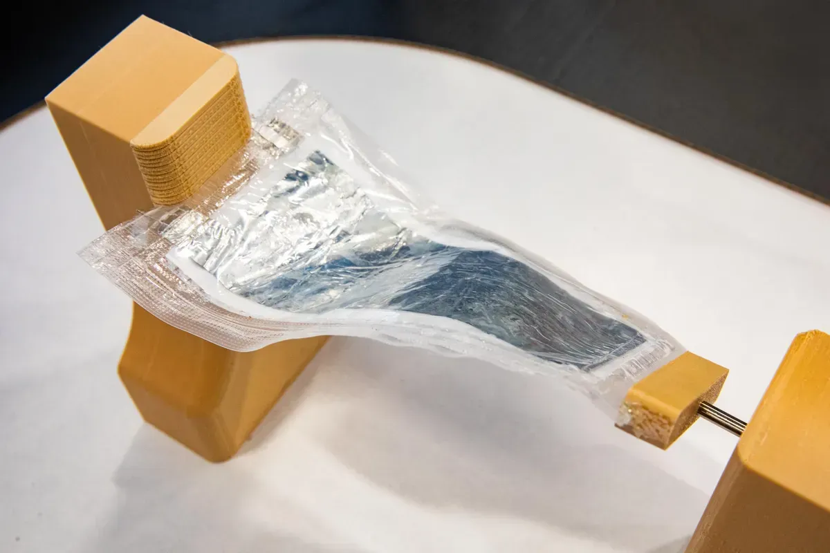 A clear plastic-wrapped object suspended between two tan wooden supports on a white surface.