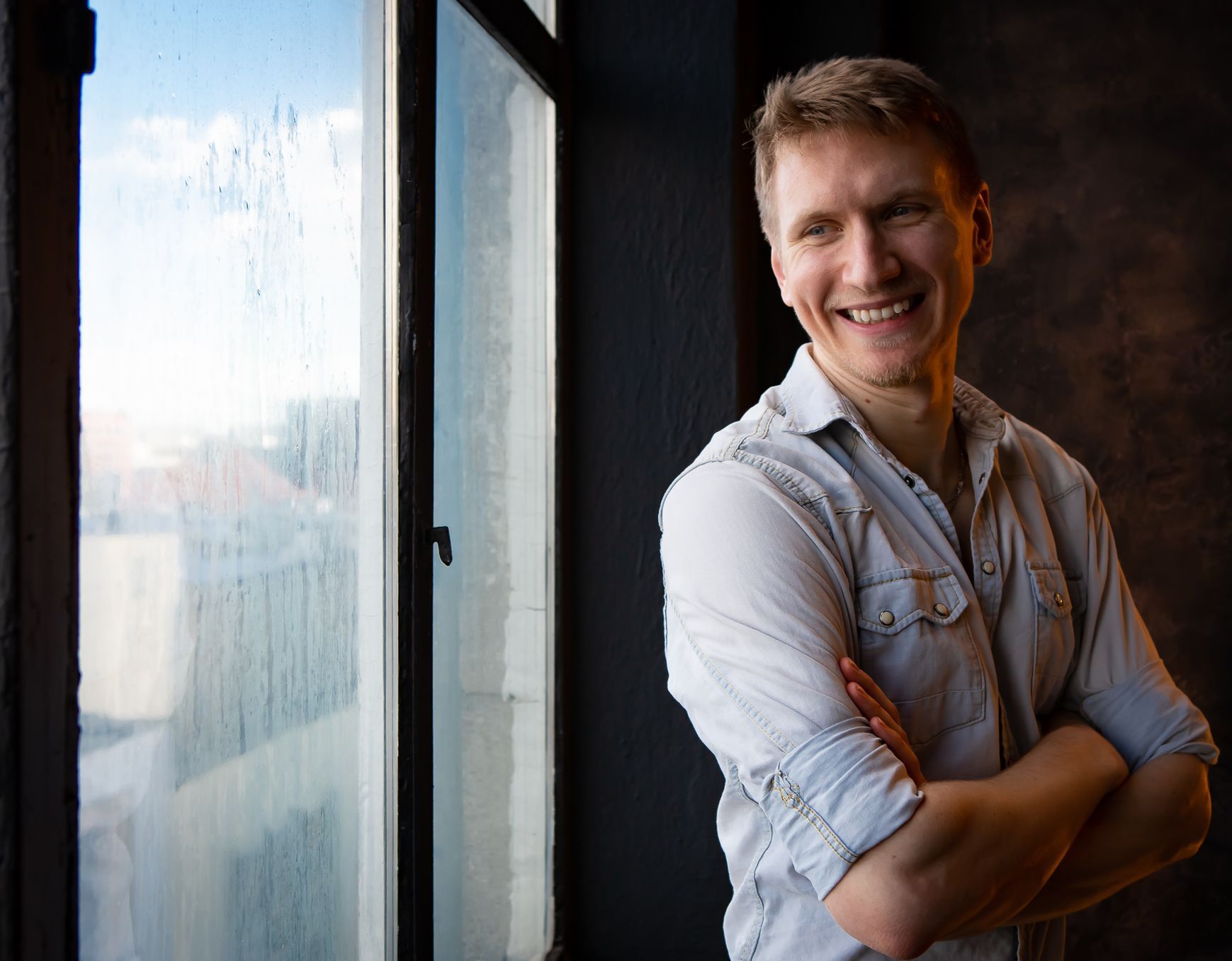 A man is standing in front of a window with his arms crossed and smiling.