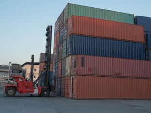A red reach stacker stands beside a towering stack of multicolored shipping containers in an industrial yard.