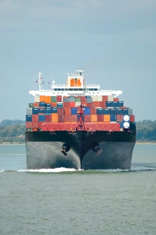 A massive, dark-hulled cargo ship loaded with colorful shipping containers moves through calm water toward the camera.
