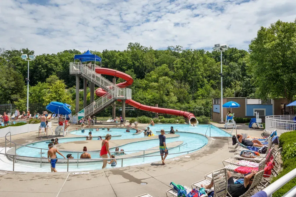 Outdoor community pool with tall red water slide and families enjoying summer activities.