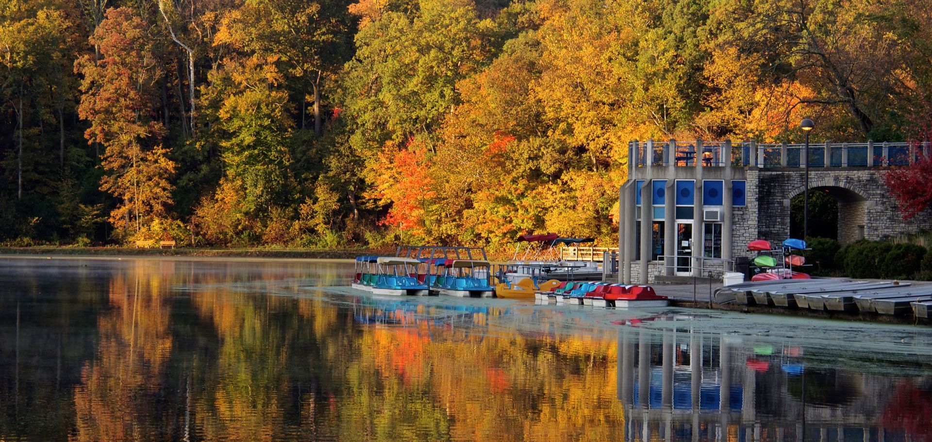 Sharon Woods lake in autumn with colorful fall foliage, boathouse, and paddleboats on the water.
