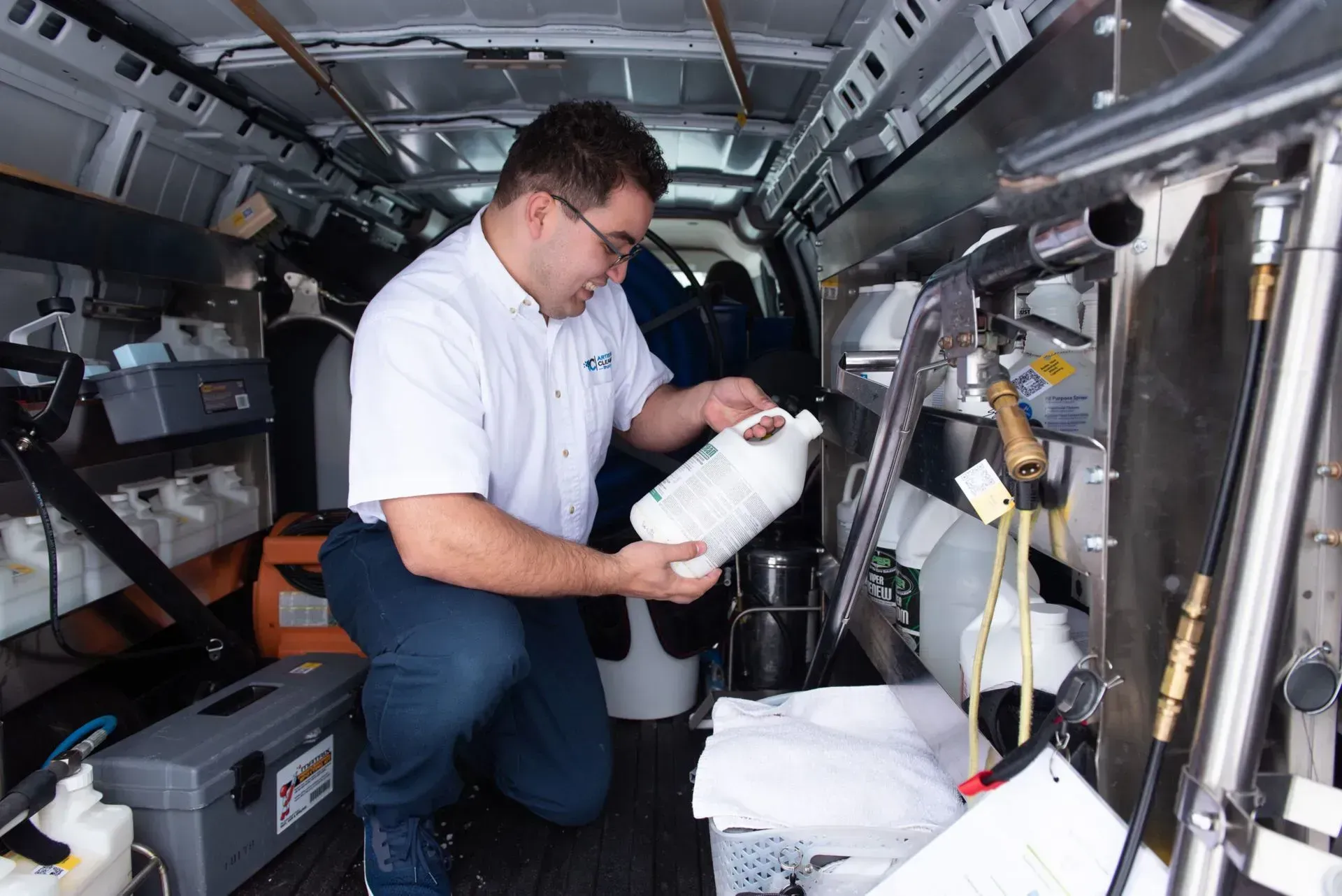 A technician in a uniform inspects a white chemical jug inside a well-organized service van.