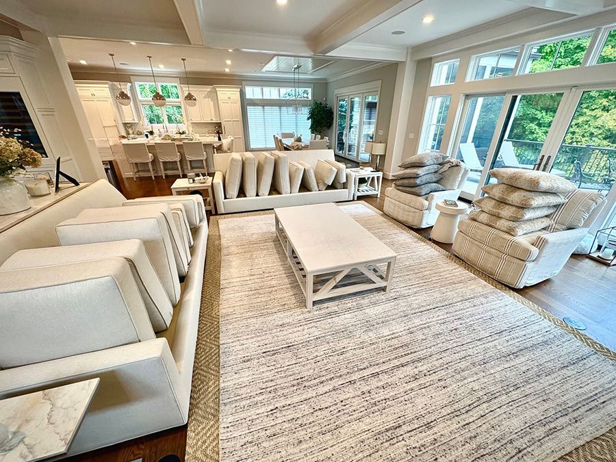 A bright, modern living room with cream sofas and armchairs arranged around a wooden coffee table on a large textured rug.