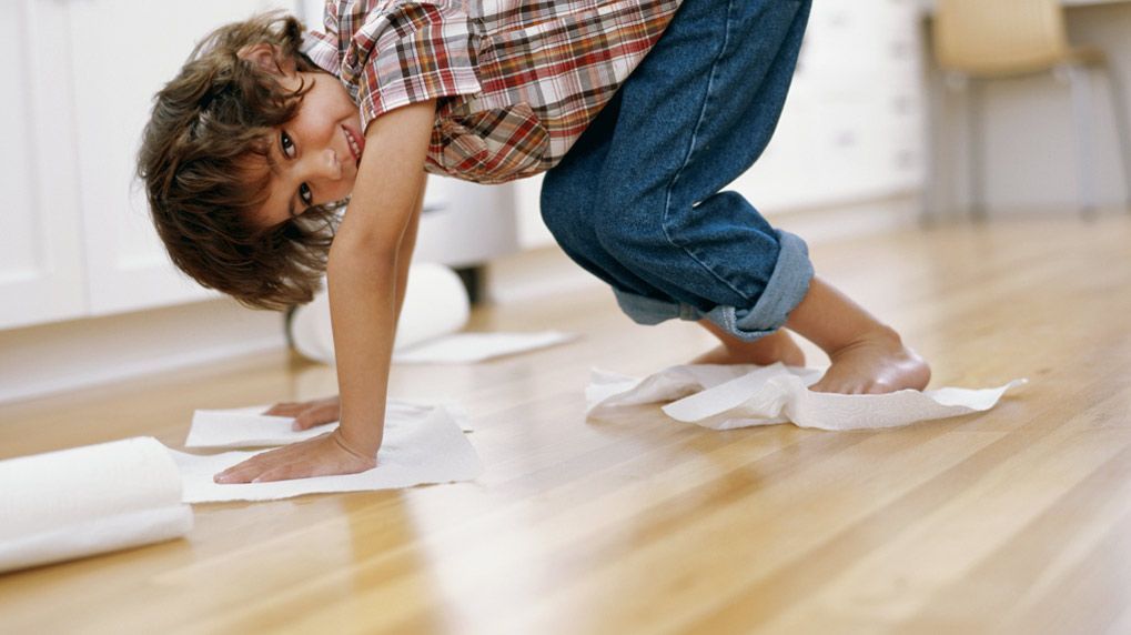 A person on hands and knees on a wooden kitchen floor, using paper towels under their hands and feet to slide.