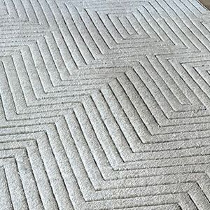 A textured, light-colored area rug featuring a repeating geometric pattern of carved, parallel zig-zag lines.