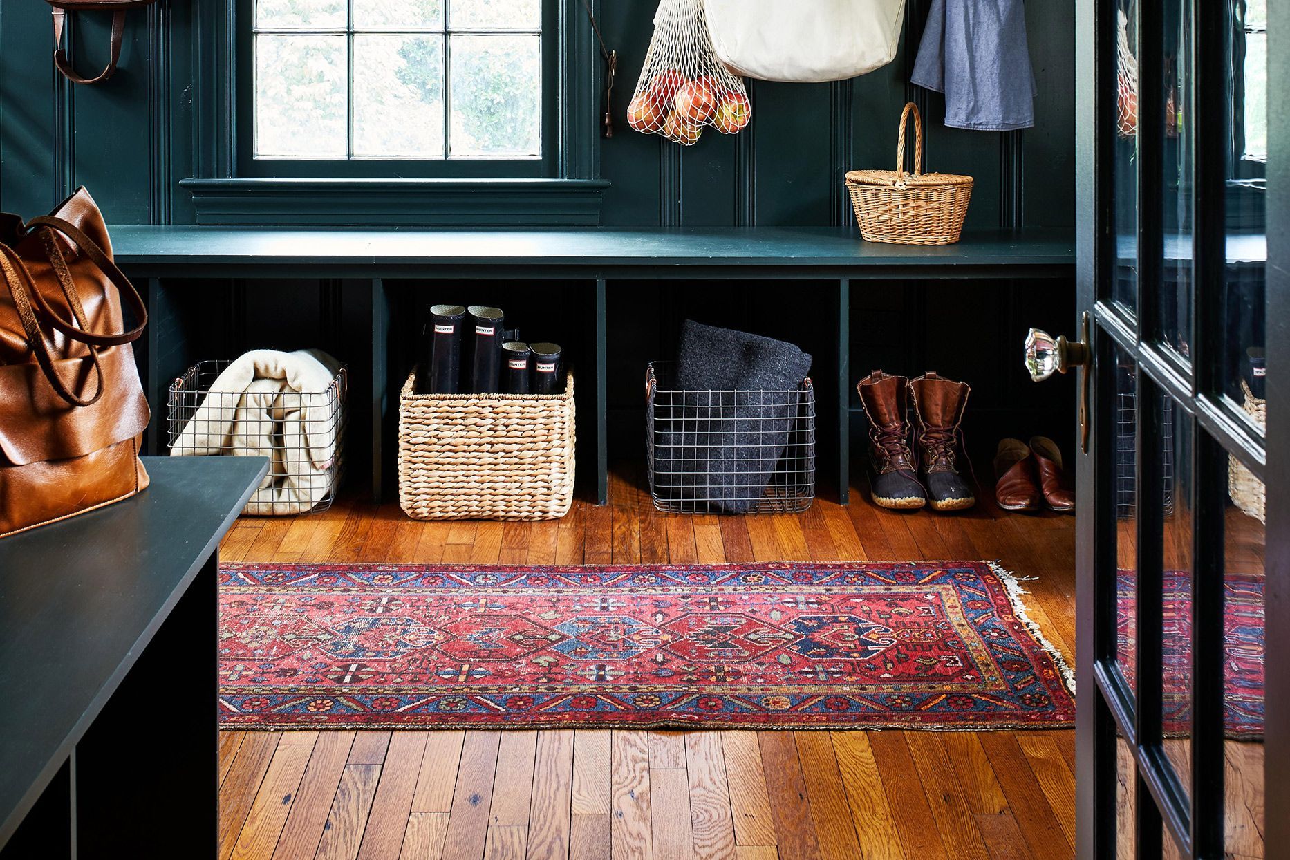 A mudroom with dark green built-in shelves, a red patterned rug on wood floors, and baskets holding shoes and gear.