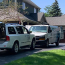 Two white work vans parked in a residential driveway with their rear doors open in front of a multi-story house.