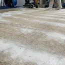 A person in protective gear operates a machine to clean a large, patterned area rug covered in cleaning foam.