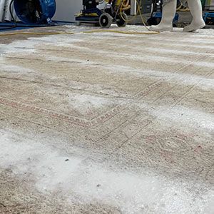 A person in protective gear operates a machine to clean a large, patterned area rug covered in cleaning foam.