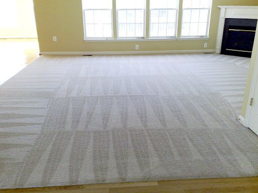 A tan carpeted living room floor with vacuum cleaner track marks, featuring a fireplace and large windows in the back.