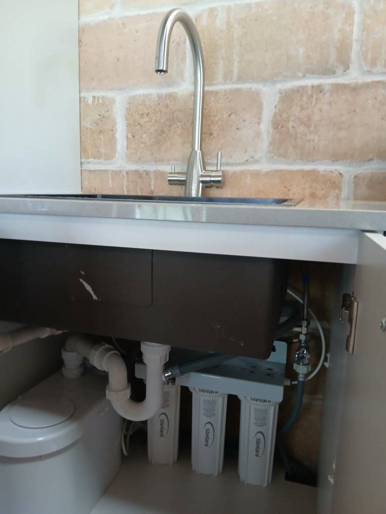 Under-sink cabinet with exposed plumbing, white filters, and a stainless sink faucet above.