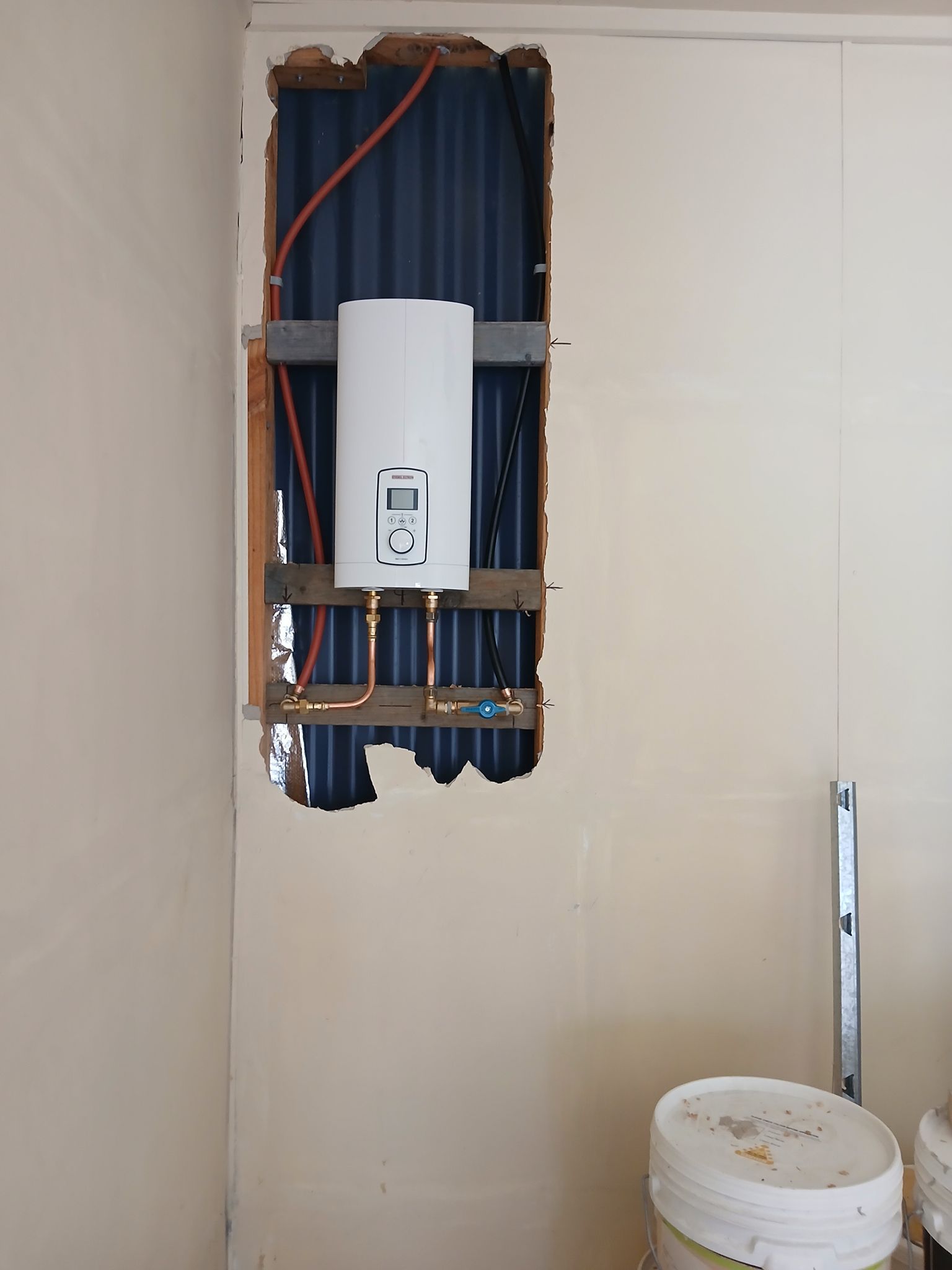 Wall-mounted white water heater in an unfinished wall cavity with exposed pipes and wiring.