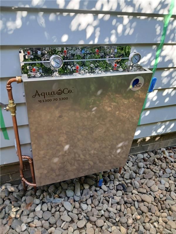 Outdoor AquaCo irrigation control box beside a house wall, with copper piping and rock landscaping