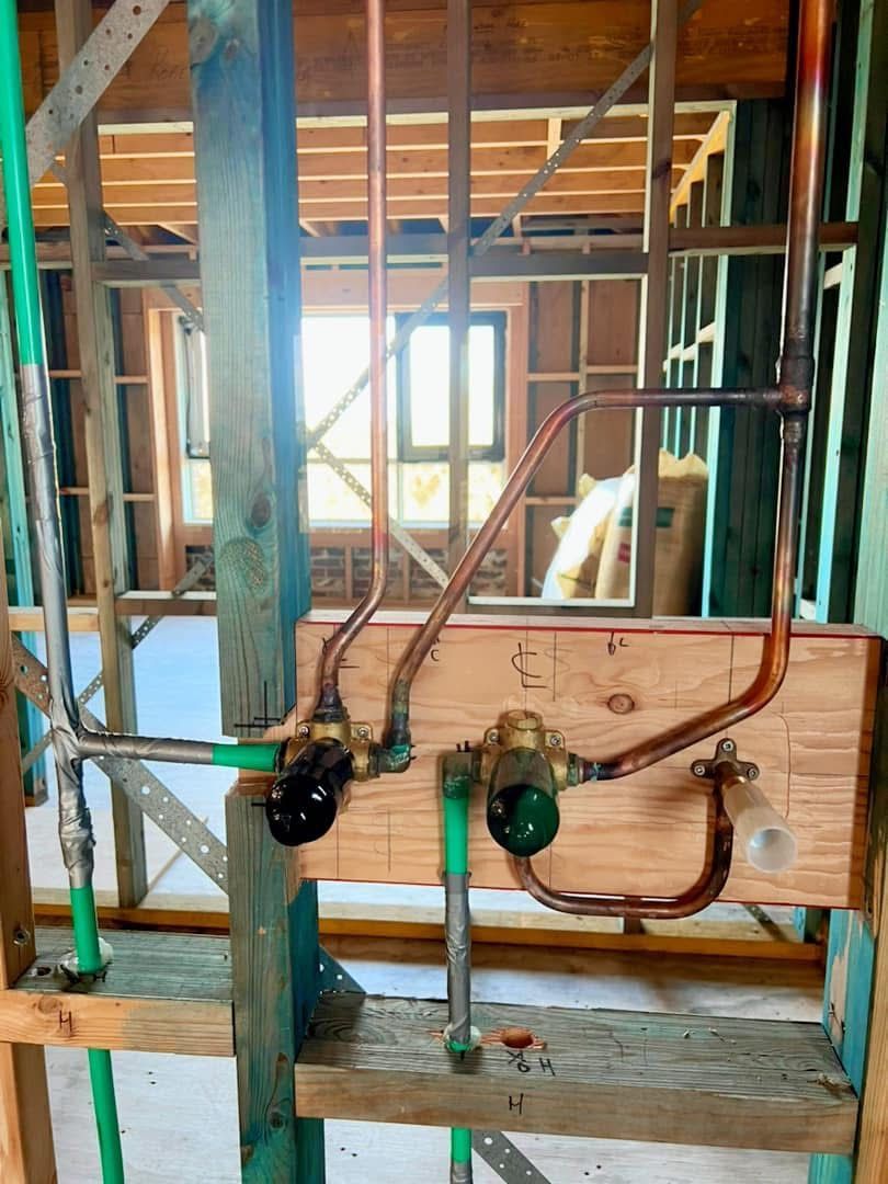 Exposed plumbing pipes and valves in a framed wall under construction, with a window in the background.