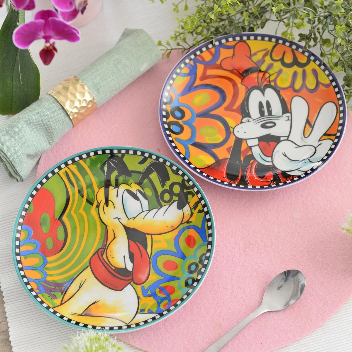 Twee kleurrijke borden met Disney-figuren, Goofy en Pluto, op een roze ondergrond, met een lepel en een servet.