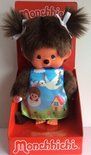 Monchhichi-pop in een doos, gekleed in een blauwgroene jurk met een cartoonachtig tafereel; bruine vacht, witte strikjes.