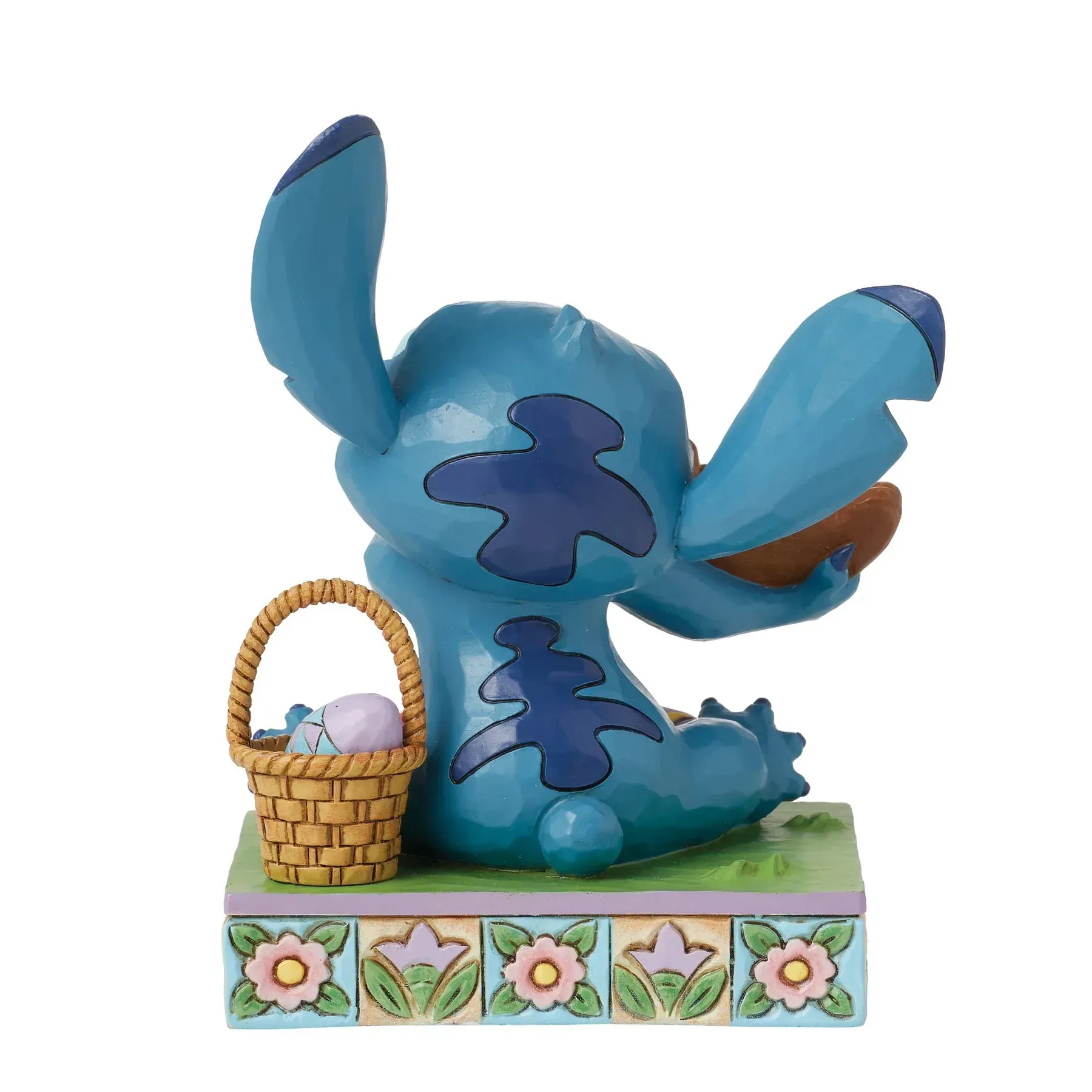 Een Stitch-figuurtje dat een chocoladekonijntje eet; paaseieren en een mandje liggen er vlakbij. Blauwe, bruine en pastelkleuren.