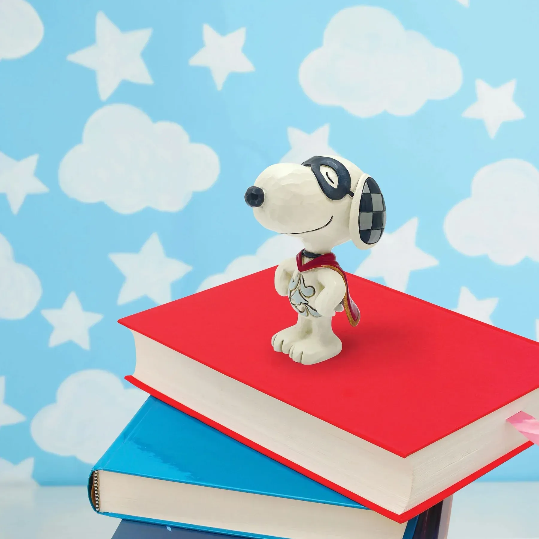 Een Snoopy-figuurtje met een bril en een sjaal staat bovenop een stapel boeken tegen een achtergrond met een hemelthema.